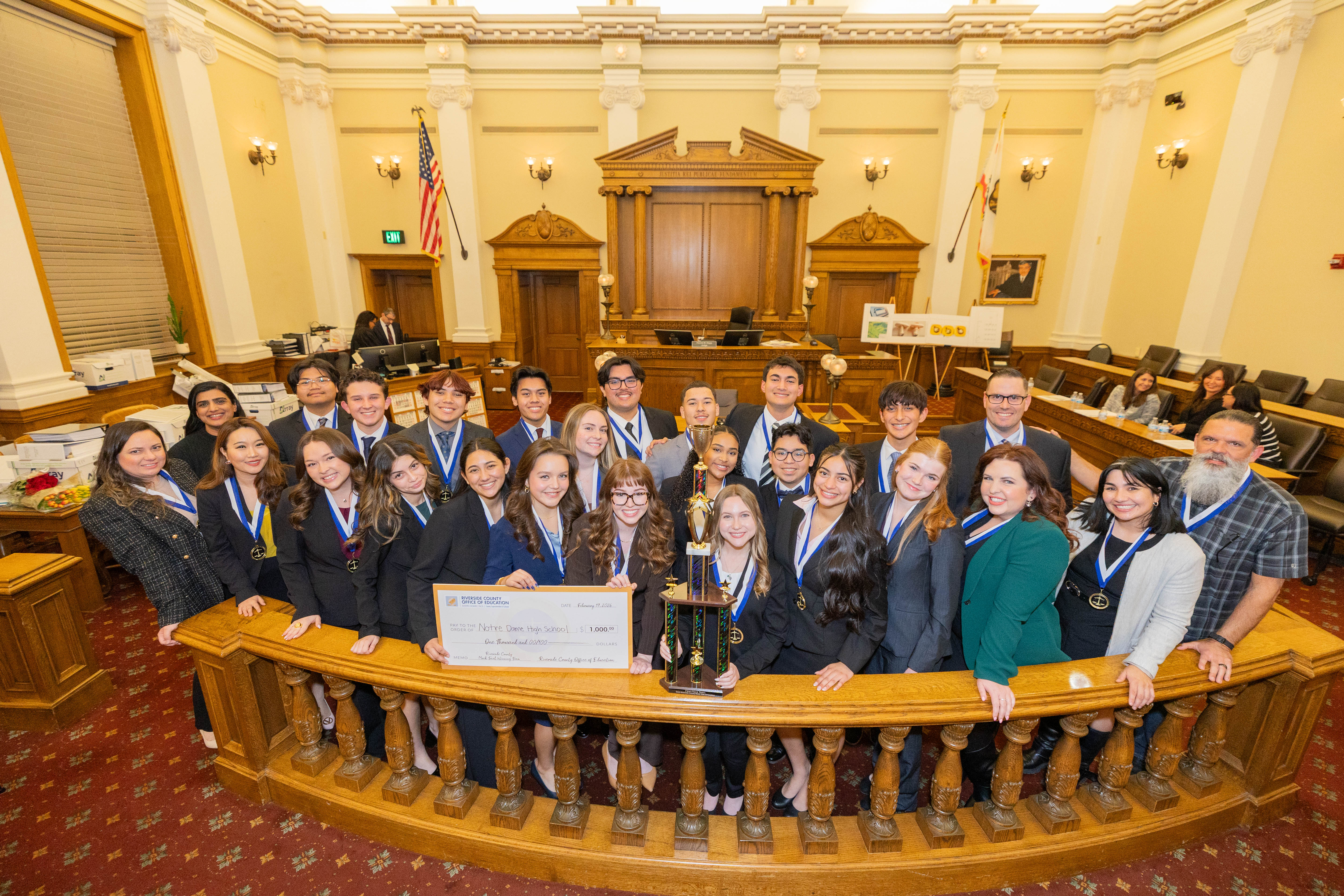Large team of students wearing suits in historic courtroom with oversized check and trophy