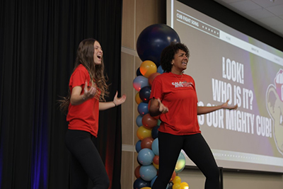 Two young adults performing on stage next to project slide that read Look! Who Is It? It's Our MIghty Cub!