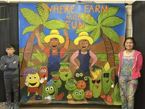 Gabby and Antonio standing side by side by their colorful poster that reads "Where Farm Meets Fun" during the 2025 Indio Date Festival