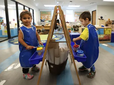 Two preschoolers wearing aprons painting on opposite sides of an easel
